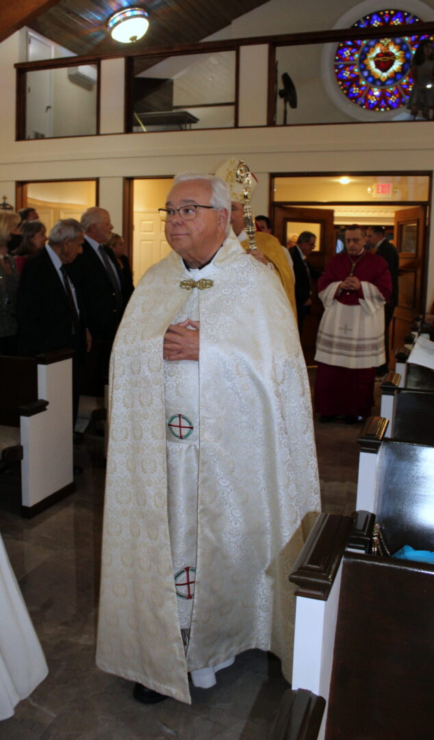 A priest with white hair and wearing an eyeglass while walking on the aisle