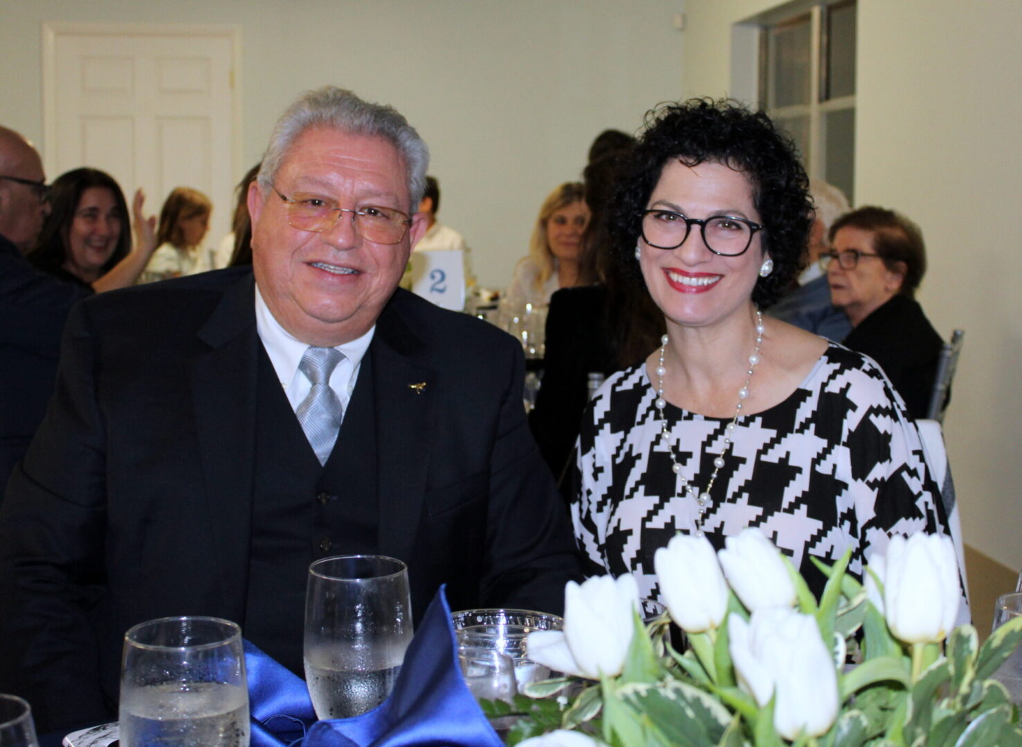 A man wearing a black suit with silver tie and a woman wearing a black-and-white top with pearl accessories