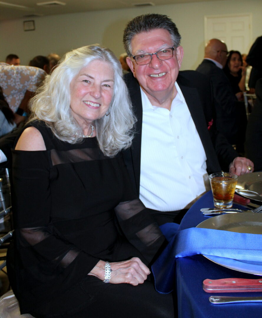A man wearing a black suit and white polo and a woman with gray hair wearing a black dress