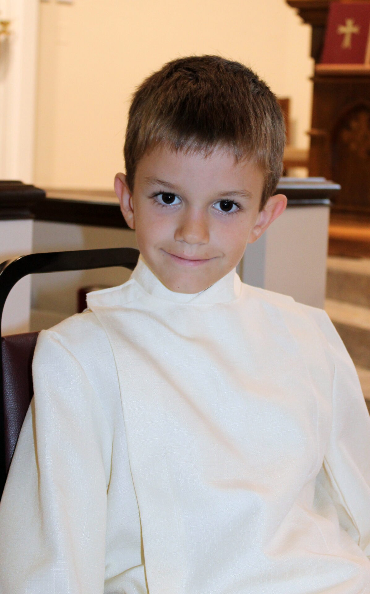 A young boy wearing a white sacristan uniform while sitting down on a brown metal chair