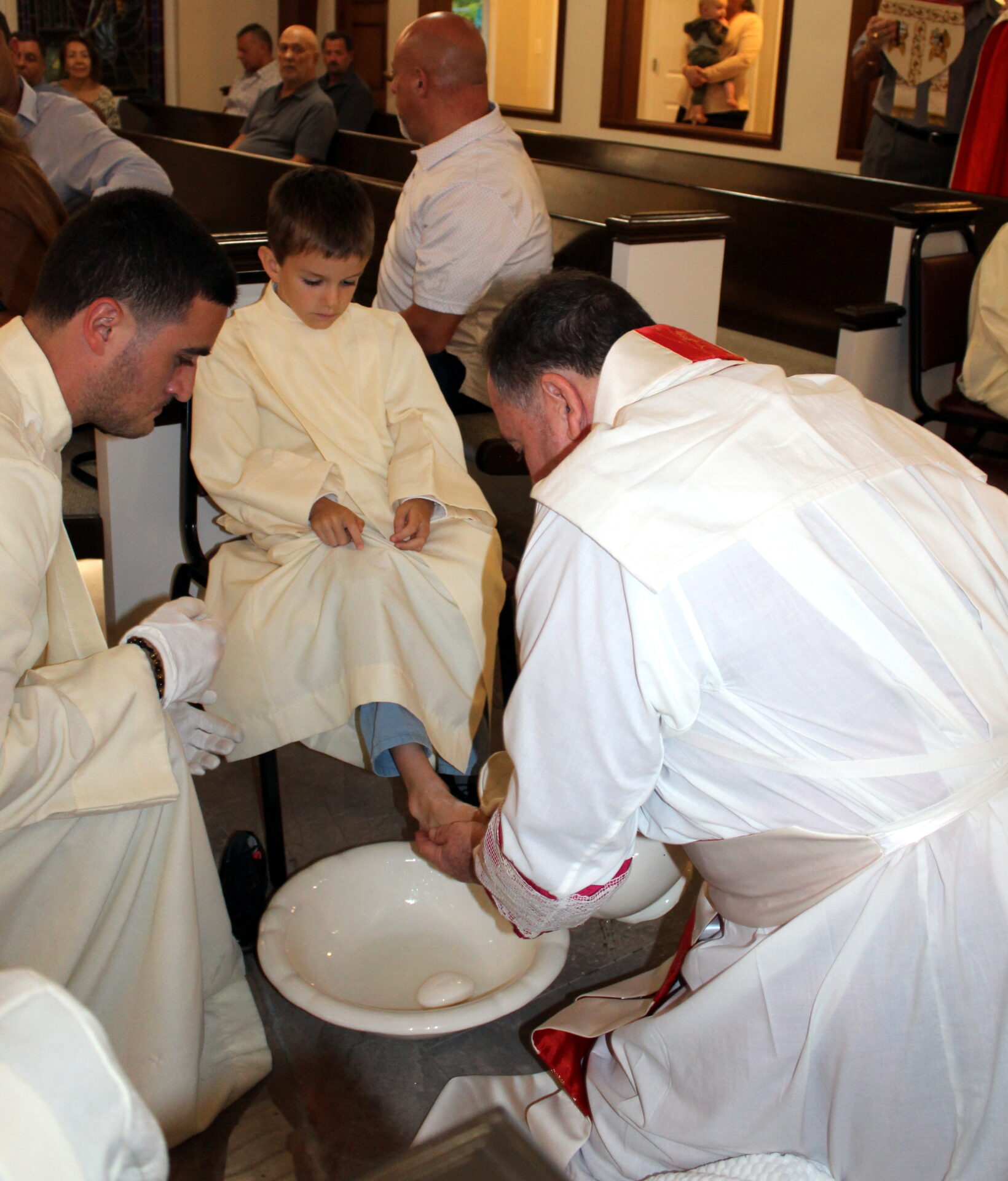 A priest washing a young sacristan’s feet beside another sacristan who’s wearing white gloves
