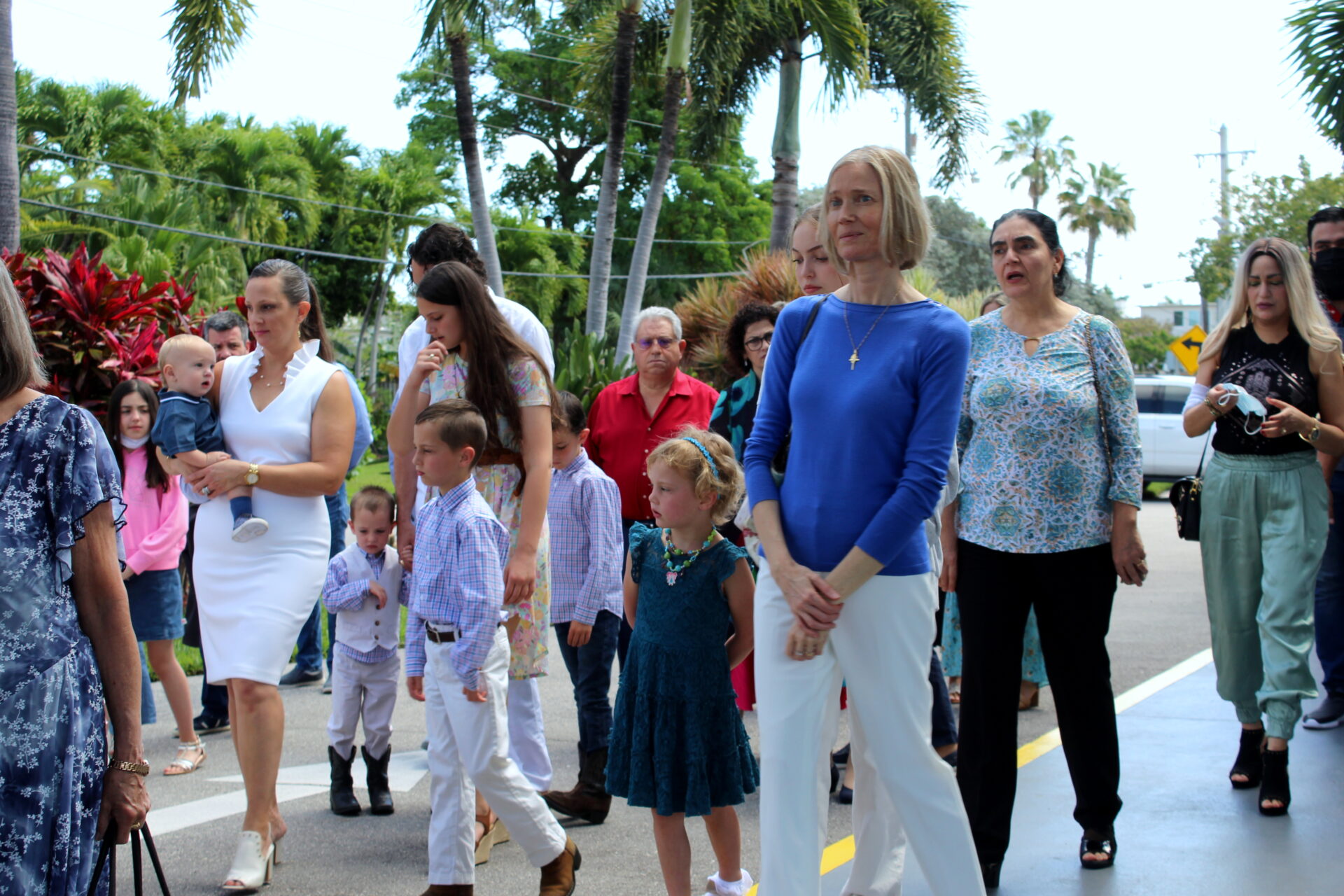 A woman with short blonde hair wearing a blue top and white pants with gold cross necklace