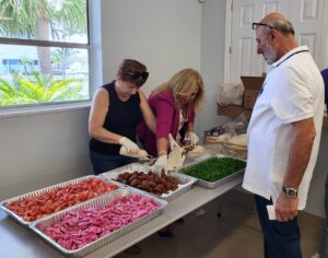 Two women preparing food for a guy wearing a white shirt with eyeglass on his forehead